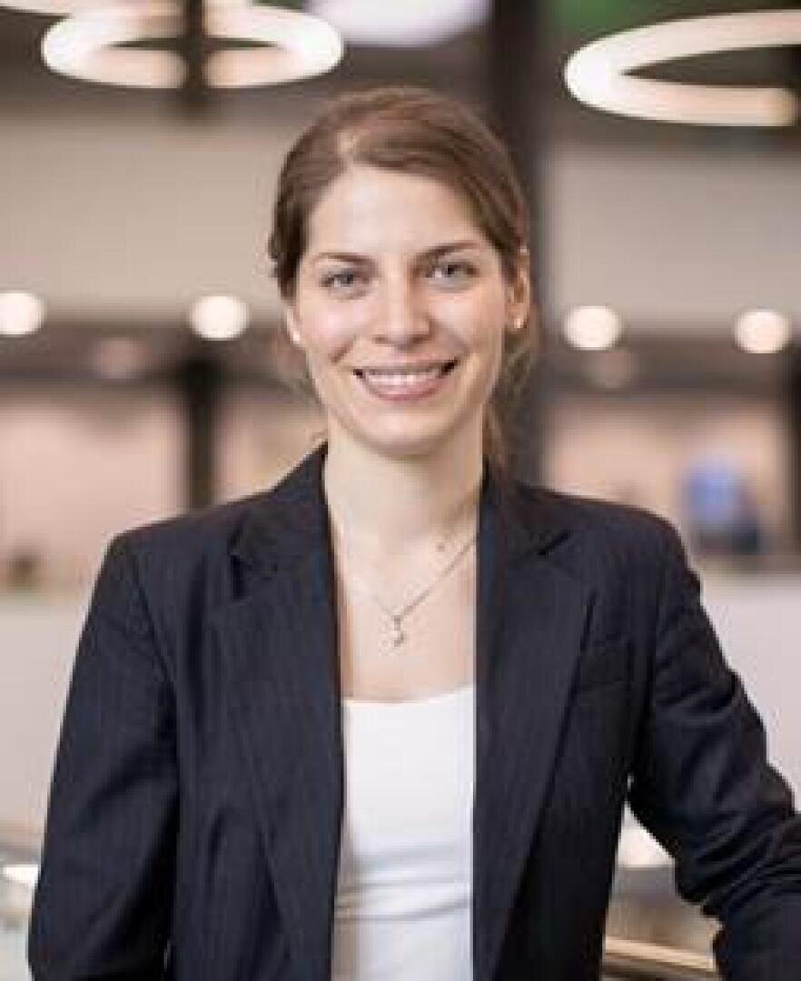 Eine Frau mit zurückgebundenem hellbraunem Haar, die einen dunklen Blazer und ein weißes Oberteil trägt, lächelt in einer modernen Inneneinrichtung mit verschwommenen Lichtern und kreisförmigen Armaturen im Hintergrund in die Kamera.