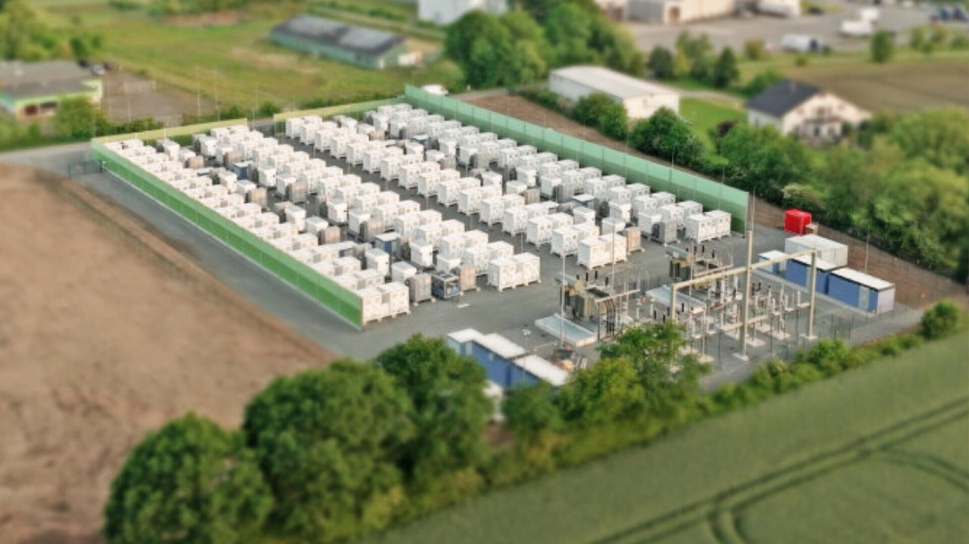 Aerial view of a fenced-in facility with rows of white containers, probably battery storage and electrical infrastructure, surrounded by trees and nearby buildings.