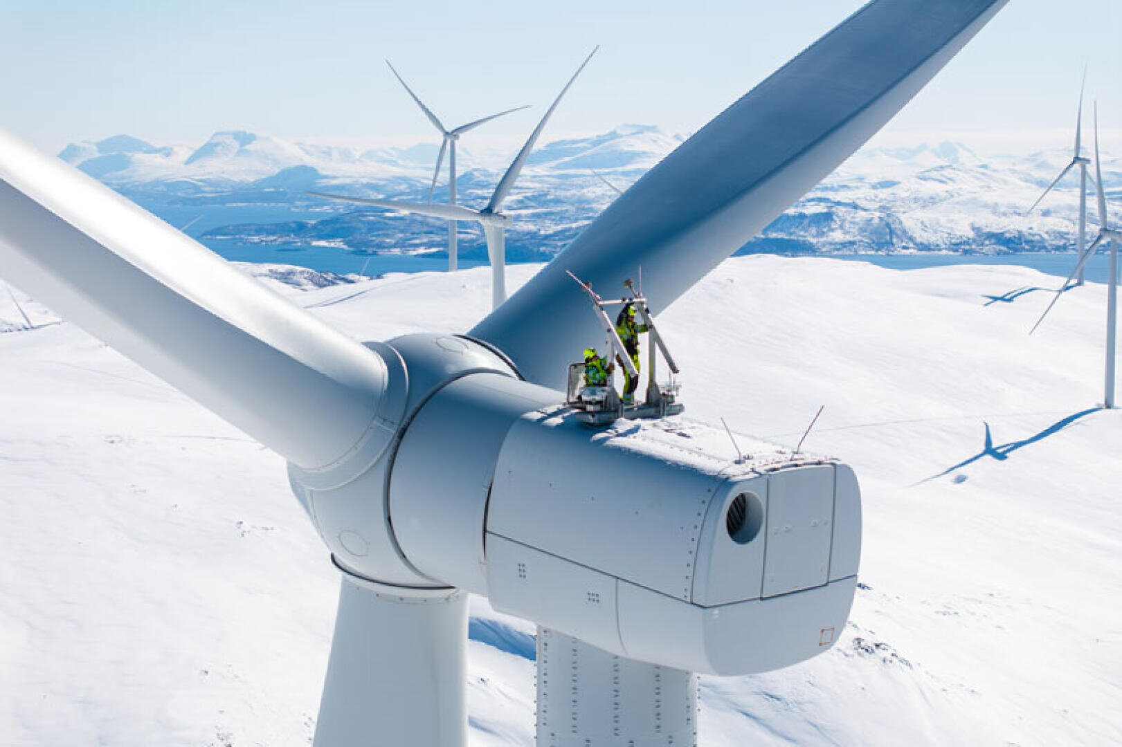 Zwei Arbeiter in Schutzausrüstung führen Wartungsarbeiten auf einer Windkraftanlage durch, umgeben von schneebedeckten Bergen und weiteren Windkraftanlagen im Hintergrund unter einem strahlend blauen Himmel.