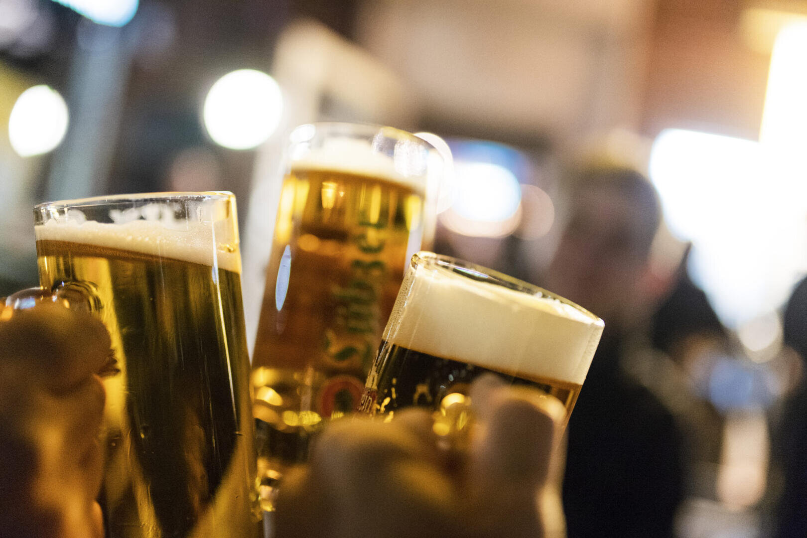 Drei Hände halten Biergläser und stoßen an. Im Hintergrund sind Menschen und warme Lichter unscharf, was auf eine lebhafte Gesellschaft oder Feier schließen lässt.