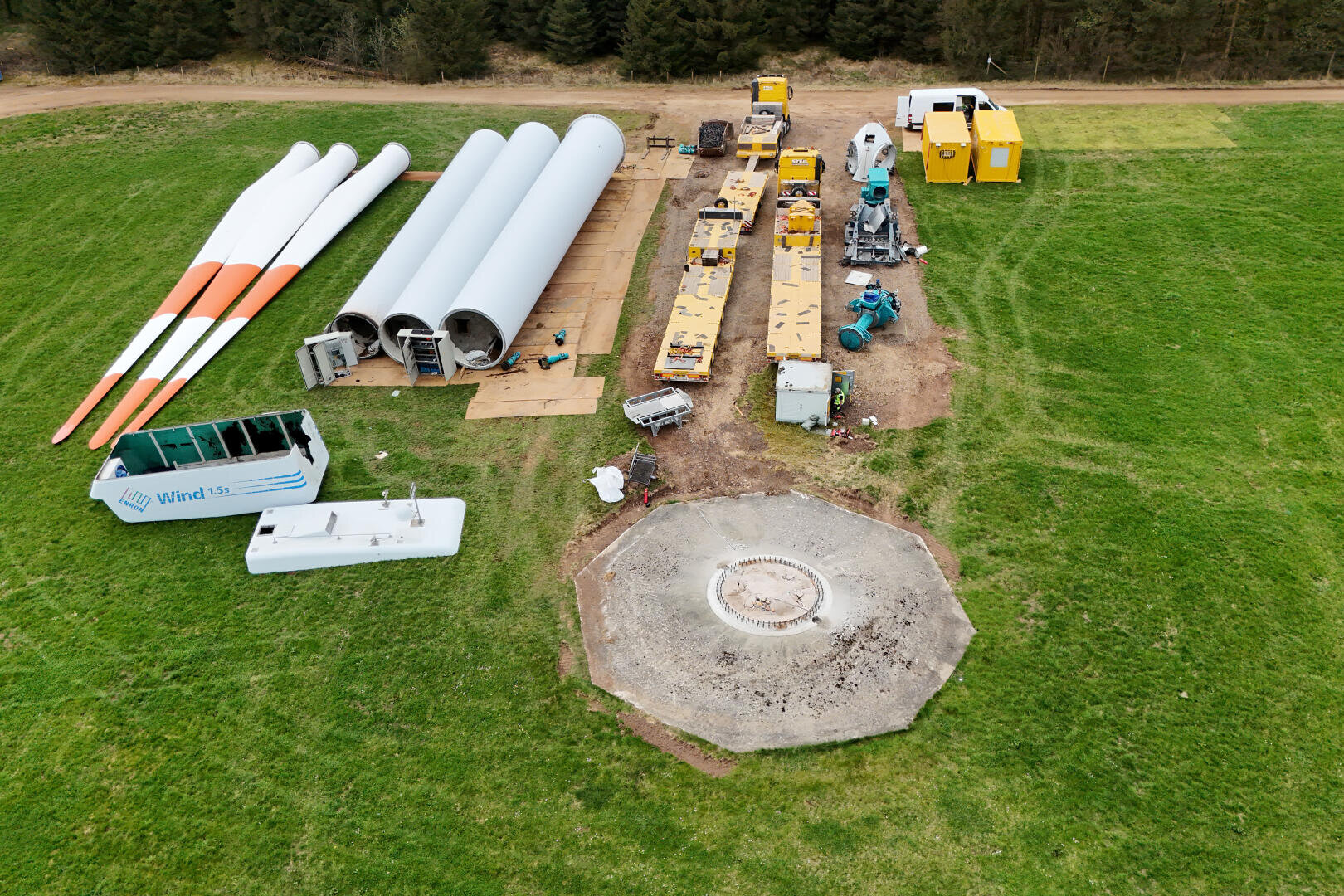 Aerial view of wind turbine parts, construction vehicles and equipment arranged in a meadow near a circular concrete foundation, with trees and a dirt road in the background.