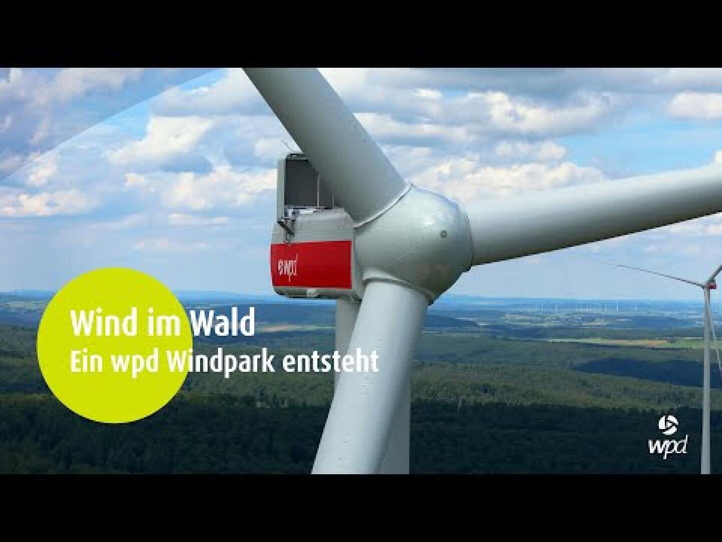 Nahaufnahme einer Windkraftanlage in einem Waldgebiet mit sanften Hügeln und Bäumen im Hintergrund. Der Text auf Deutsch lautet: „Wind im Wald. Ein wpd Windpark entsteht.“ und das wpd-Logo ist sichtbar.