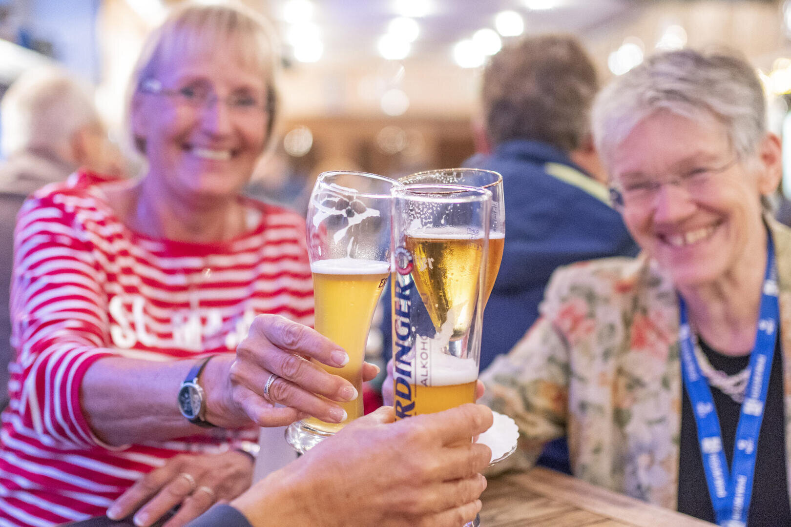 Zwei lächelnde ältere Frauen sitzen an einem Tisch und stoßen fröhlich mit großen Biergläsern an, während im Hintergrund eine lebhafte Innenatmosphäre herrscht.