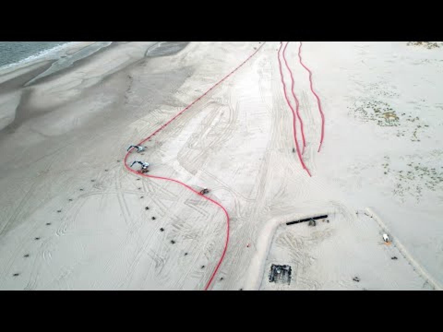 Luftaufnahme eines Sandstrandes mit mehreren langen roten Rohren, die quer über den Sand verlegt sind. In der Nähe der Rohre sind Baufahrzeuge und -geräte zu sehen, und Reifenspuren bedecken das Gebiet.