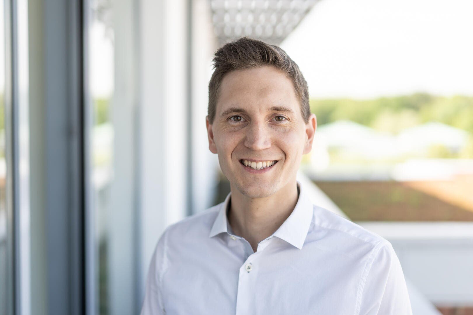 Ein lächelnder Mann mit kurzen braunen Haaren und weißem Button-up-Hemd steht draußen auf einem Balkon mit verschwommenem Grün im Hintergrund.