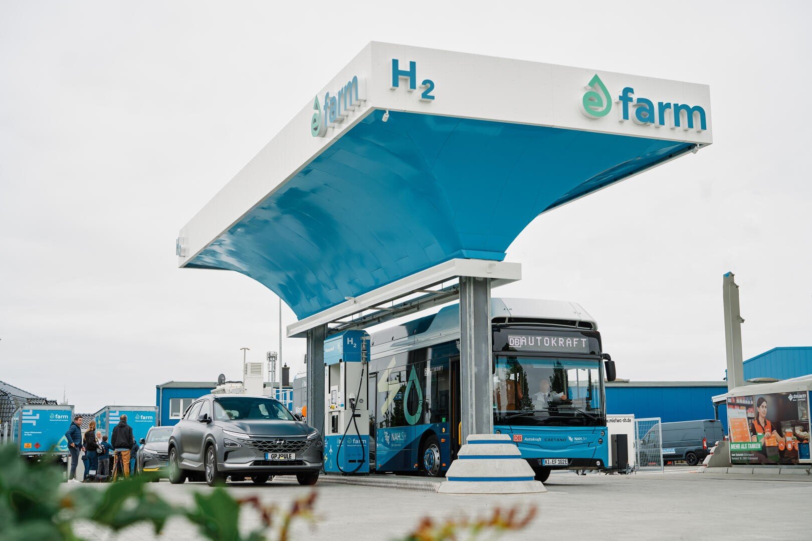 A hydrogen filling station with a blue and white canopy bearing the words e farm and H2. A bus and a car are being refueled while people stand nearby. Buildings and equipment can be seen in the background.