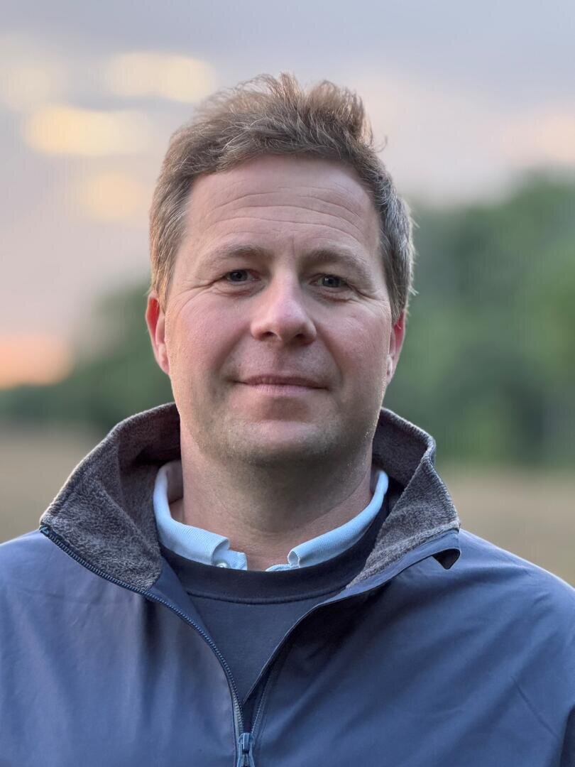 Ein Mann mit kurzen braunen Haaren, blauer Jacke und weißem Hemd mit Kragen steht im Freien, vor einem unscharfen Hintergrund aus Grünpflanzen und einem sanften Sonnenuntergang.