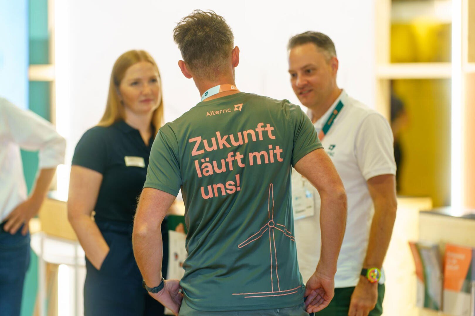 A man wearing a shirt with the German text Zukunft läuft mit uns! and a wind turbine graphic talks to two other people at an indoor event. The atmosphere appears friendly and professional.