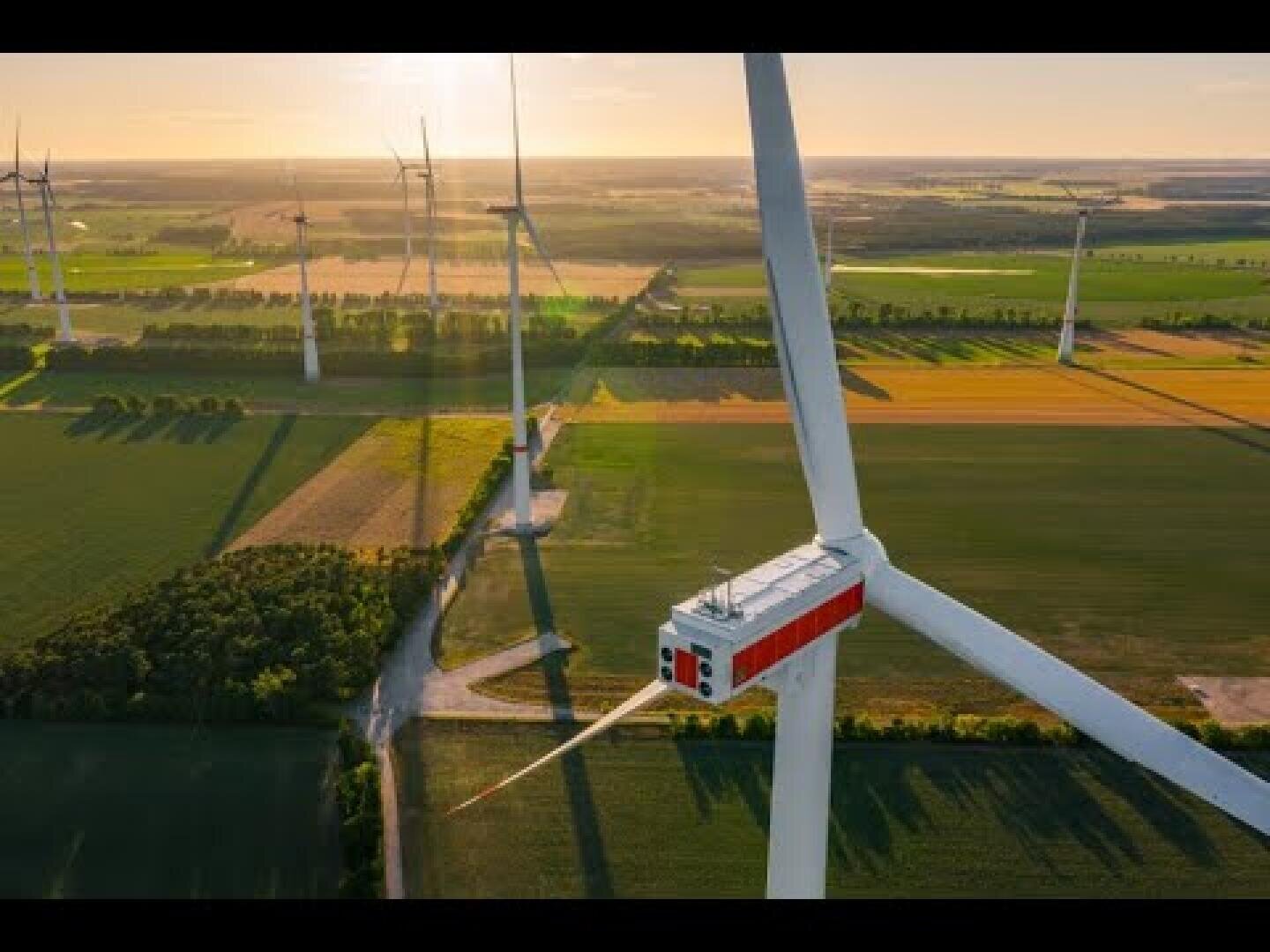 Luftaufnahme mehrerer Windturbinen auf grünem Ackerland unter klarem Himmel bei Sonnenuntergang, der lange Schatten auf die Felder wirft.