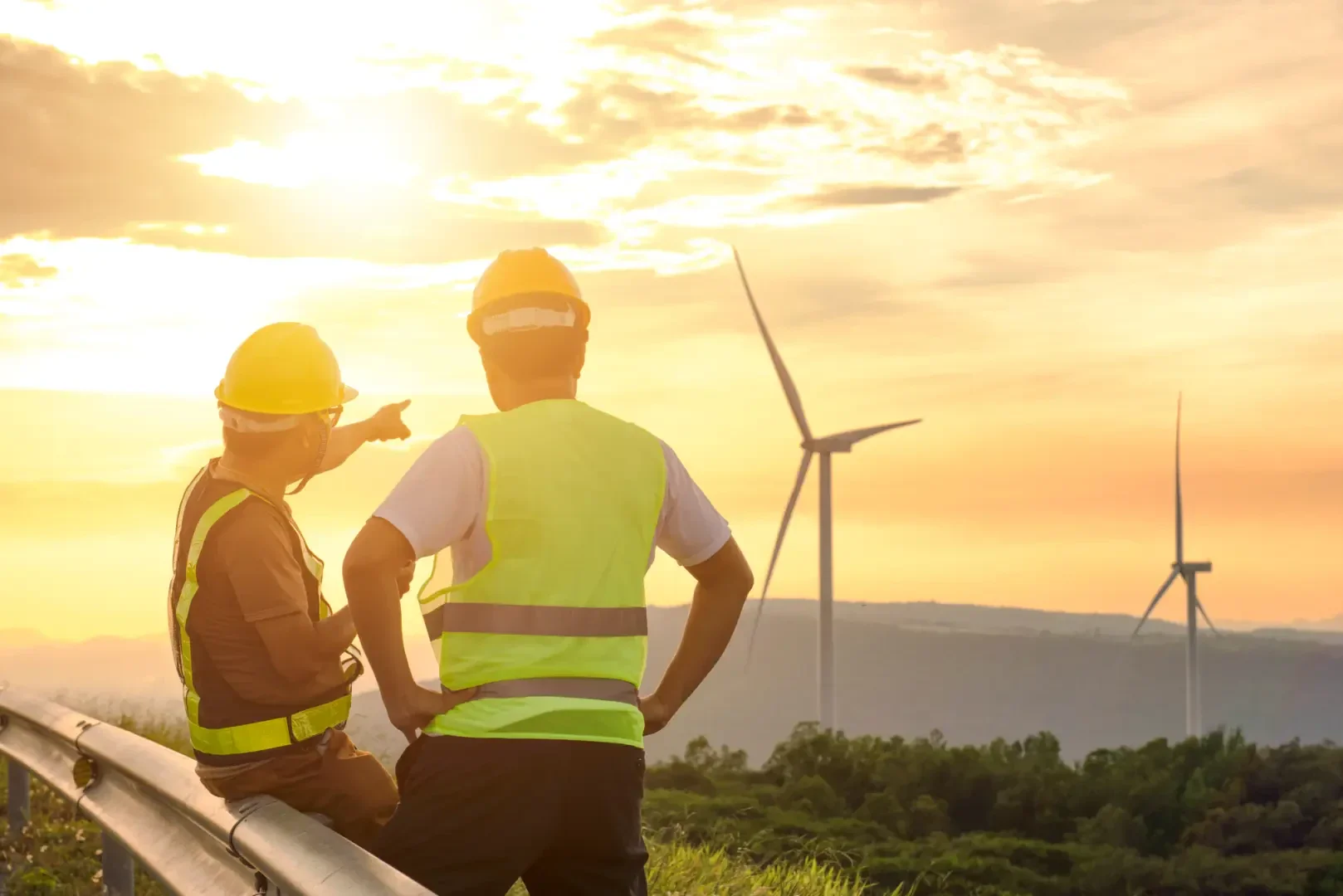 Two workers wearing hard hats and reflective vests stand near a guardrail, looking at wind turbines on a hill during sunset. One worker is pointing toward the turbines, with the sky glowing orange and yellow.
