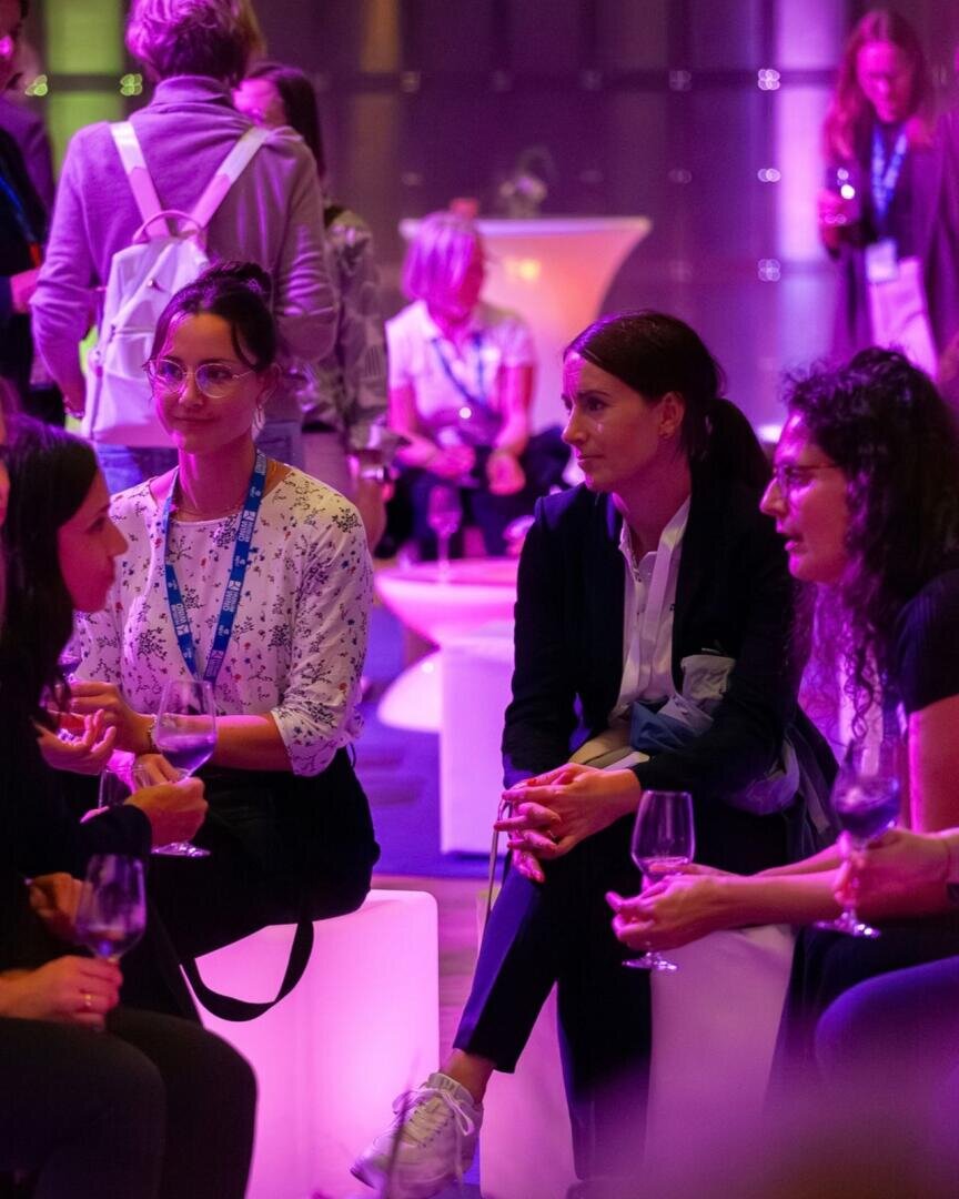 Eine Gruppe von Frauen sitzt beieinander, unterhält sich und hält Weingläser in der Hand – bei einer Veranstaltung in einem Innenraum mit violetter Beleuchtung. Im Hintergrund sind weitere Personen zu sehen, von denen einige stehen und sich unterhalten.
