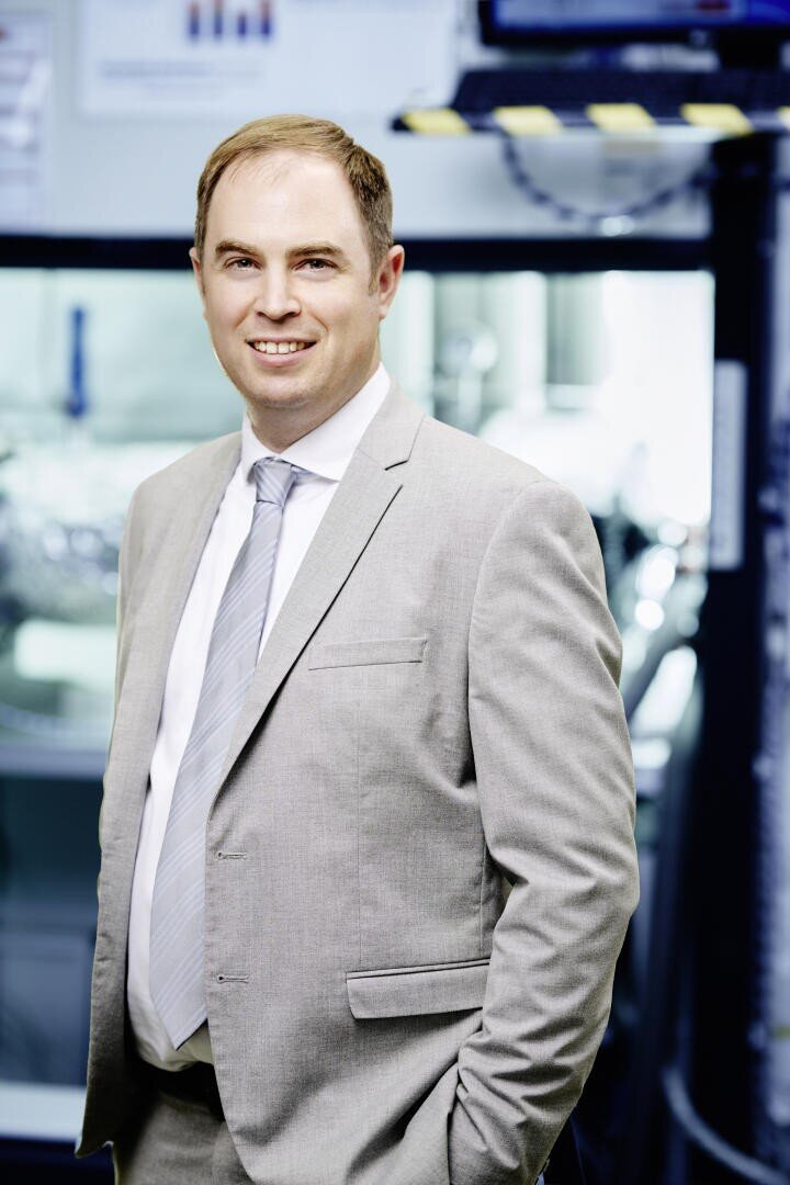 Ein Mann in einem hellgrauen Anzug mit Krawatte steht in einem Haus und lächelt in die Kamera. Der Hintergrund ist leicht unscharf und zeigt Geräte und Monitore, was auf eine professionelle oder industrielle Umgebung hindeutet.