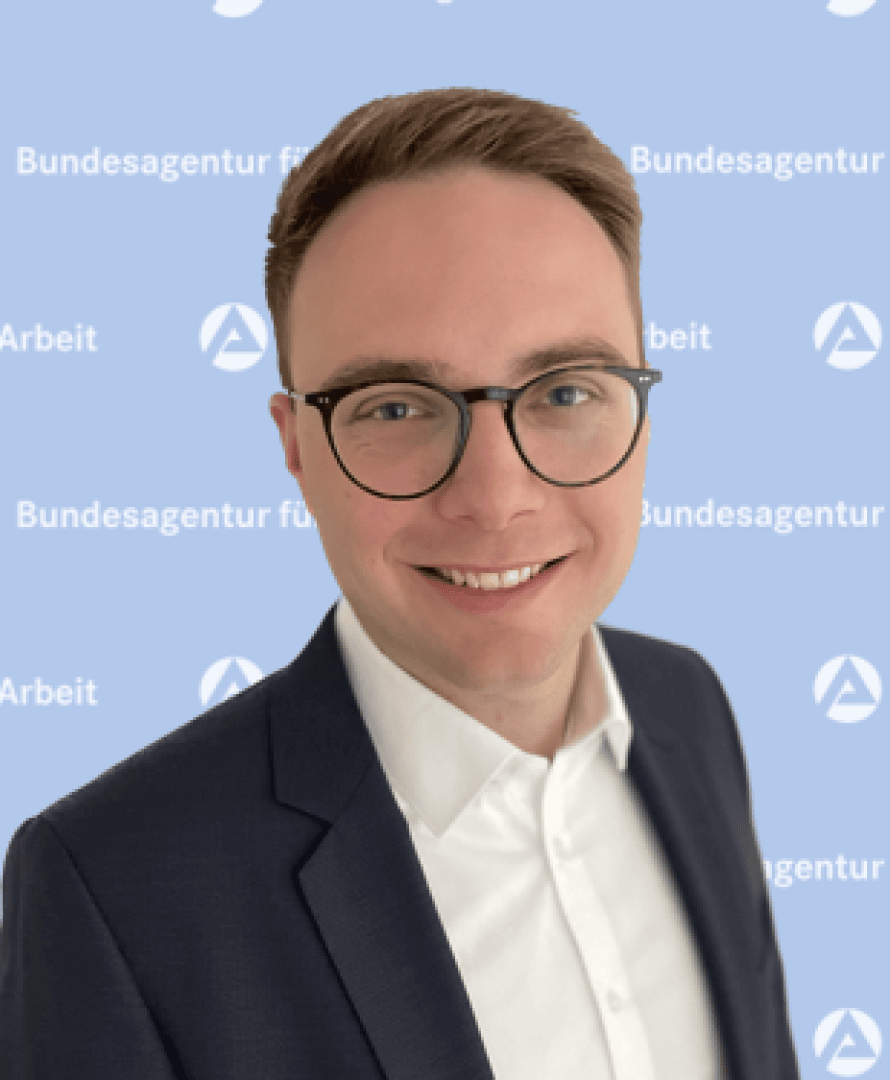 A smiling man with short brown hair, wearing round glasses, a white shirt, and a dark suit jacket stands in front of a blue background with Bundesagentur für Arbeit and its logo repeatedly displayed.