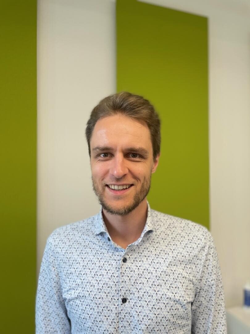 Ein Mann mit kurzen braunen Haaren und einem Bart, der ein helles gemustertes Hemd trägt, steht lächelnd vor einer Wand mit zwei großen vertikalen grünen Tafeln. Der Hintergrund ist hell und leicht unscharf.