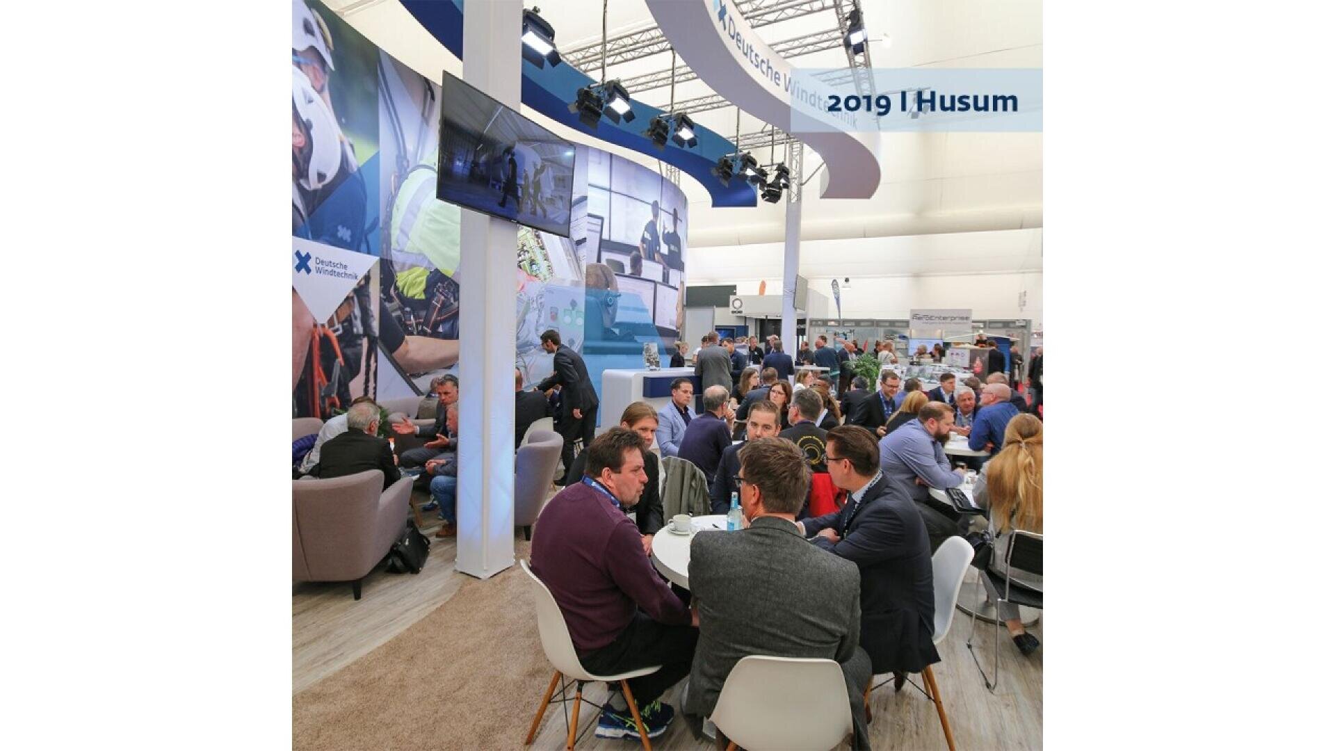 A busy exhibition booth at the 2019 Husum event, with groups of people in business attire seated at tables, networking and talking. Large display screens, promotional banners, and informational materials are visible in the background.