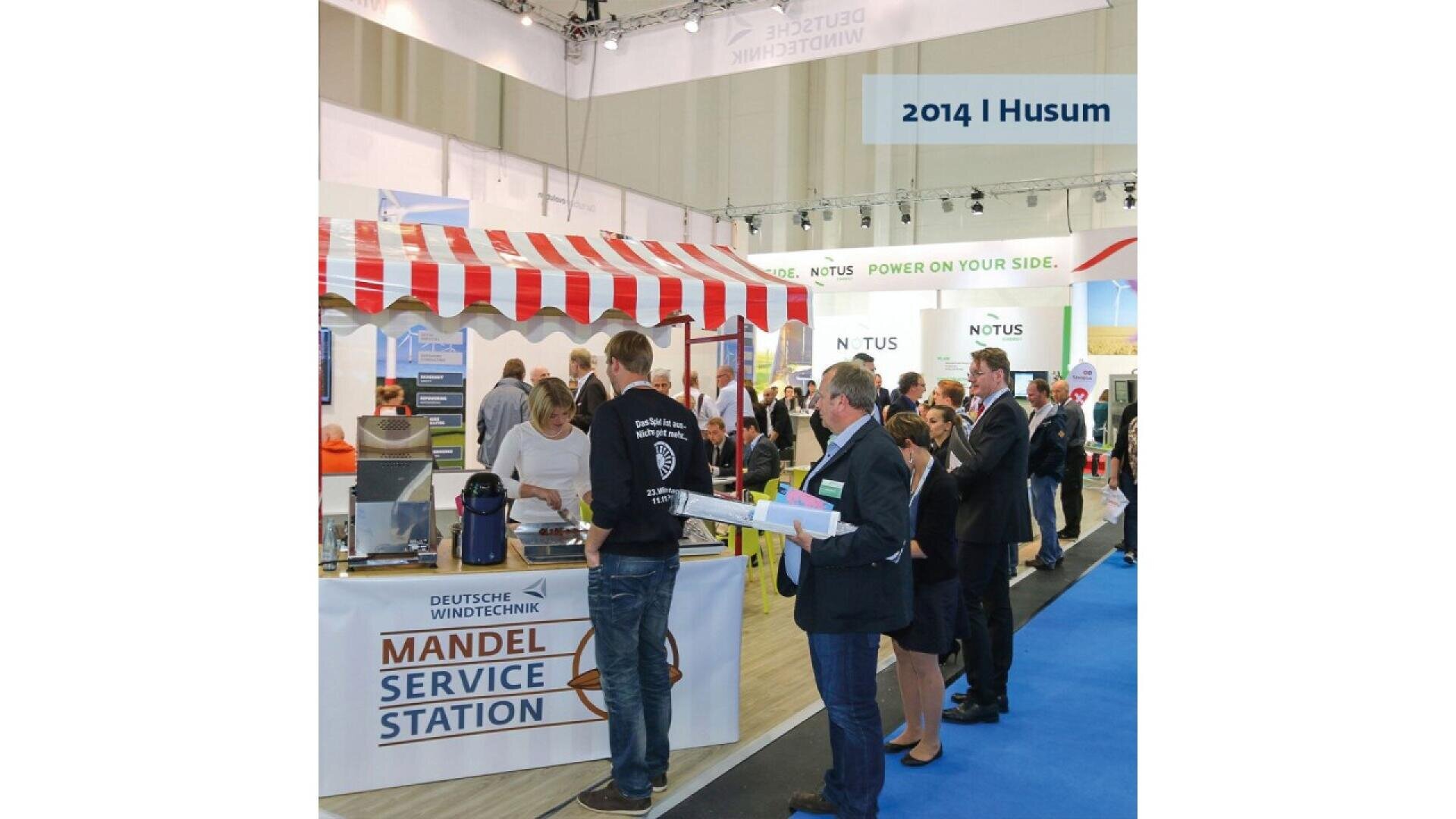 People stand in line at a booth labeled Mandel Service Station at an indoor event. The booth has a red-and-white striped canopy. A blue carpet runs alongside the booth. A sign above reads 2014 | Husum.