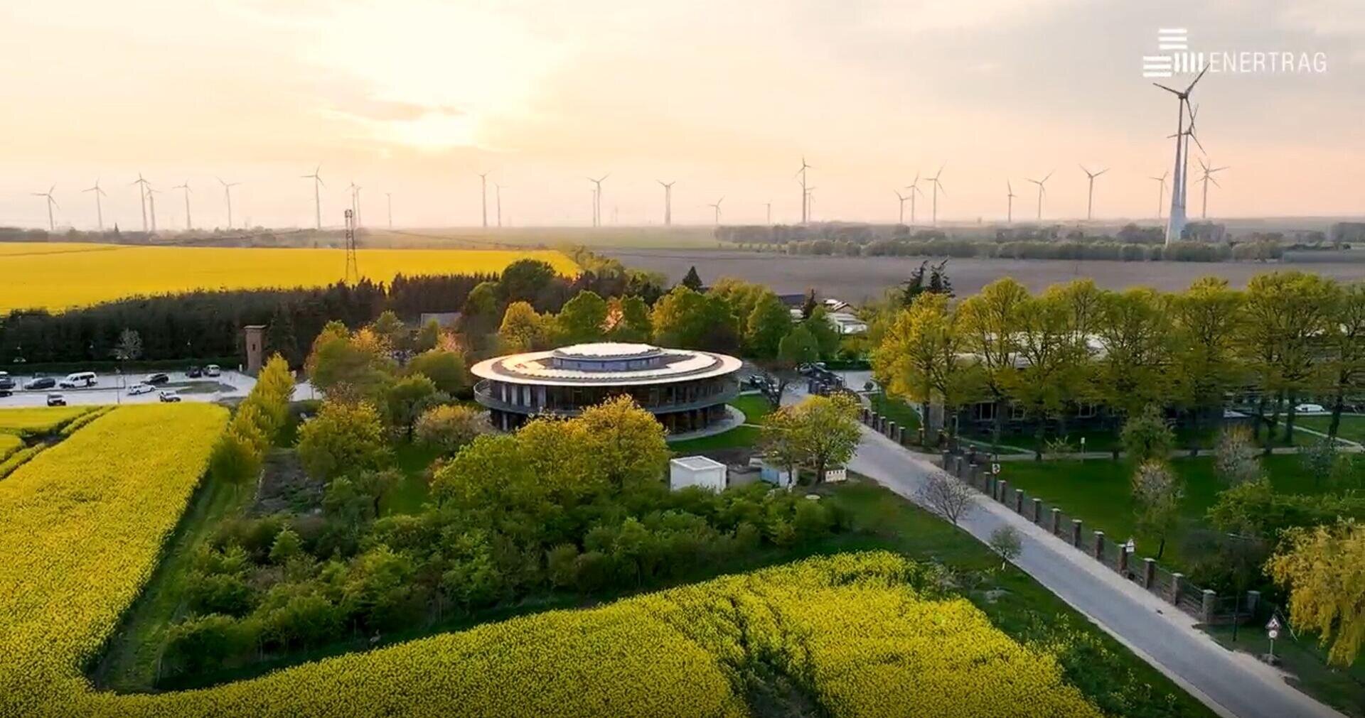 Luftaufnahme eines runden Gebäudes, das von Bäumen und gelben Feldern umgeben ist, mit Windturbinen im Hintergrund unter einem Sonnenuntergangshimmel. Mehrere Autos sind in der Nähe des Gebäudes geparkt. Das Logo "ENERTRAG" befindet sich in der oberen rechten Ecke.