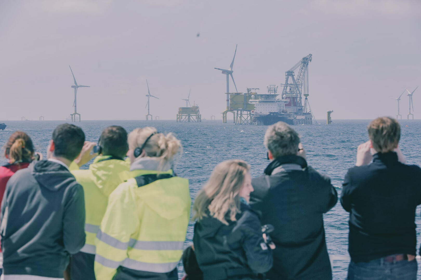 Eine Gruppe von Menschen in hellen Jacken steht am Wasser und beobachtet und fotografiert Offshore-Windkraftanlagen und Industrieplattformen in der Ferne unter einem bewölkten Himmel.