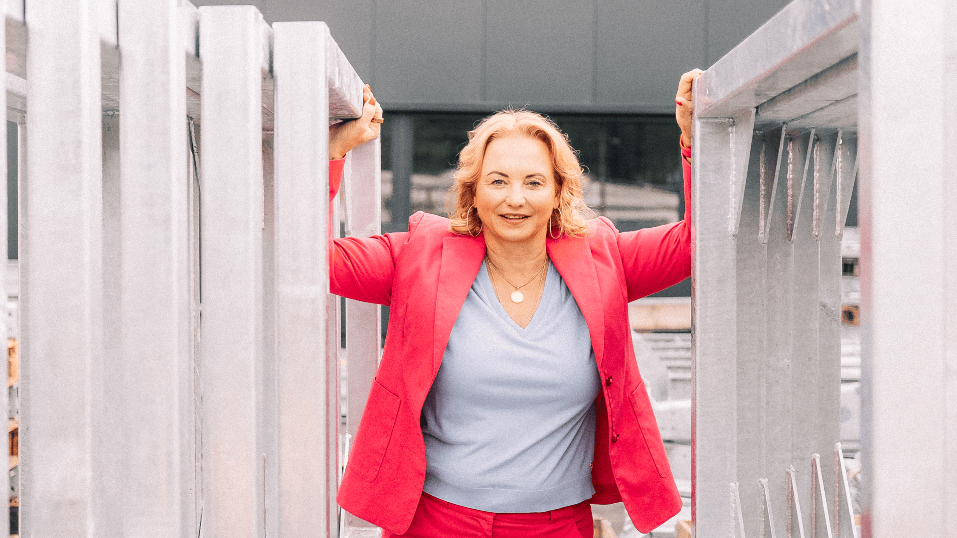 Eine Frau in einem leuchtend rosafarbenen Anzug und hellblauem Oberteil steht selbstbewusst im Freien und hält sich an zwei großen Metallträgern auf beiden Seiten fest, während im Hintergrund ein modernes Gebäude zu sehen ist.