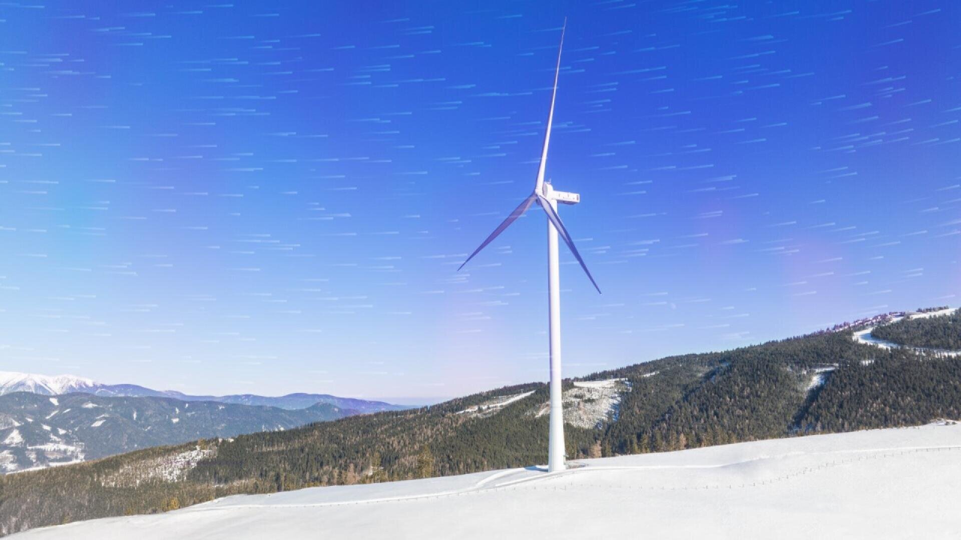 A single wind turbine stands on a snowy hillside under a clear night sky with star trails, surrounded by forested mountains in the background.