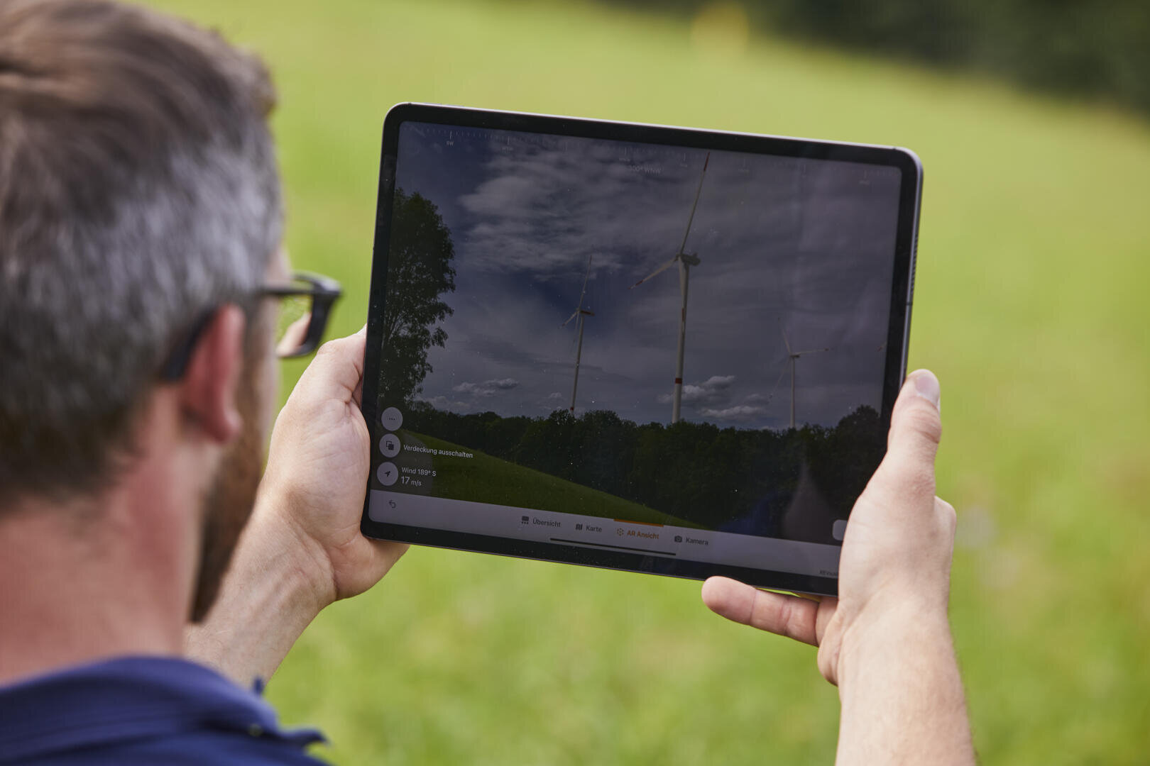 A person holding a tablet outdoors displays an image of wind turbines in a grassy landscape, with trees and a partly cloudy sky in the background.