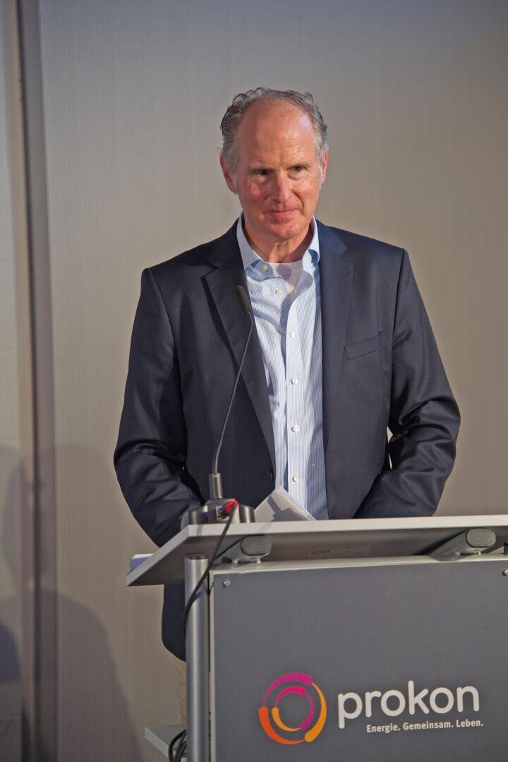 Ein Mann im Anzug steht an einem Podium mit dem prokon-Logo, hält Papiere in der Hand und spricht vor einem schlichten Hintergrund.
