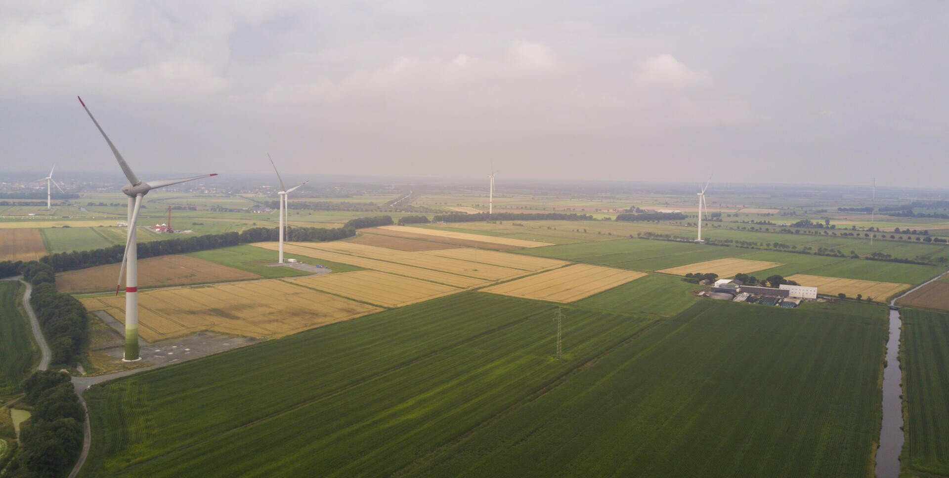Aerial view of green and yellow farmland with several large wind turbines scattered across the fields under a cloudy sky. Some buildings can be seen near the right edge of the picture.