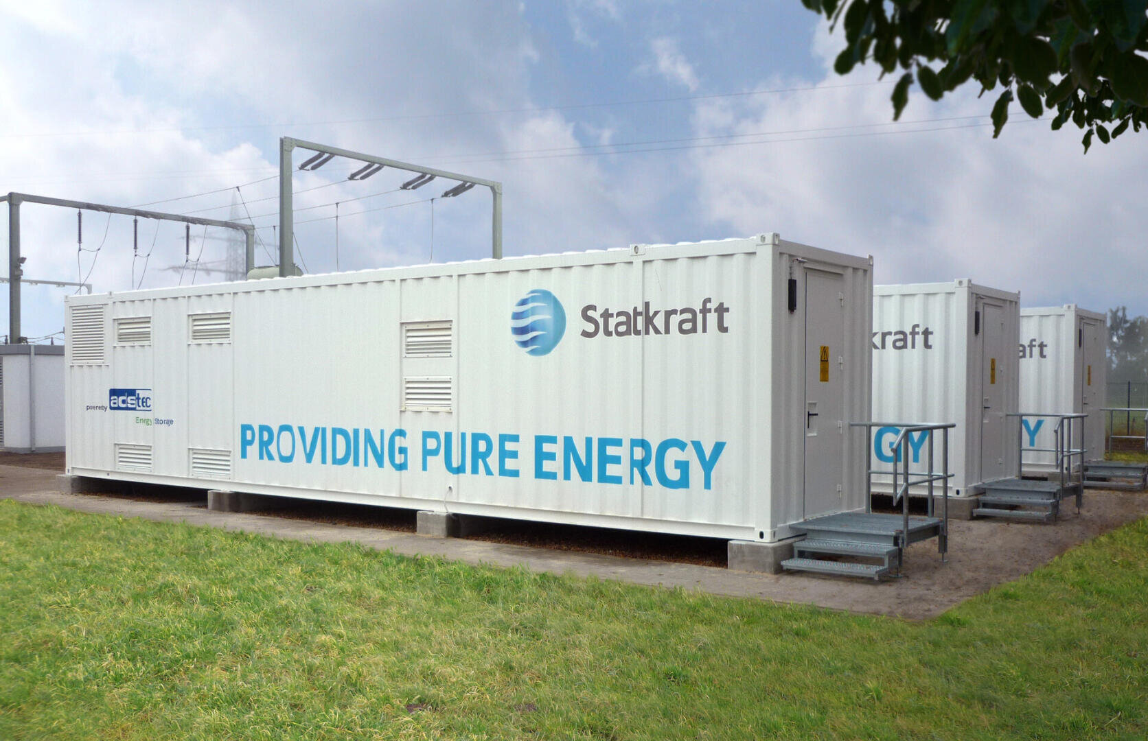 Three large white shipping containers with the inscriptions Stakraft and PROVIDING PURE ENERGY are standing under a cloudy sky on a meadow near electrical appliances.