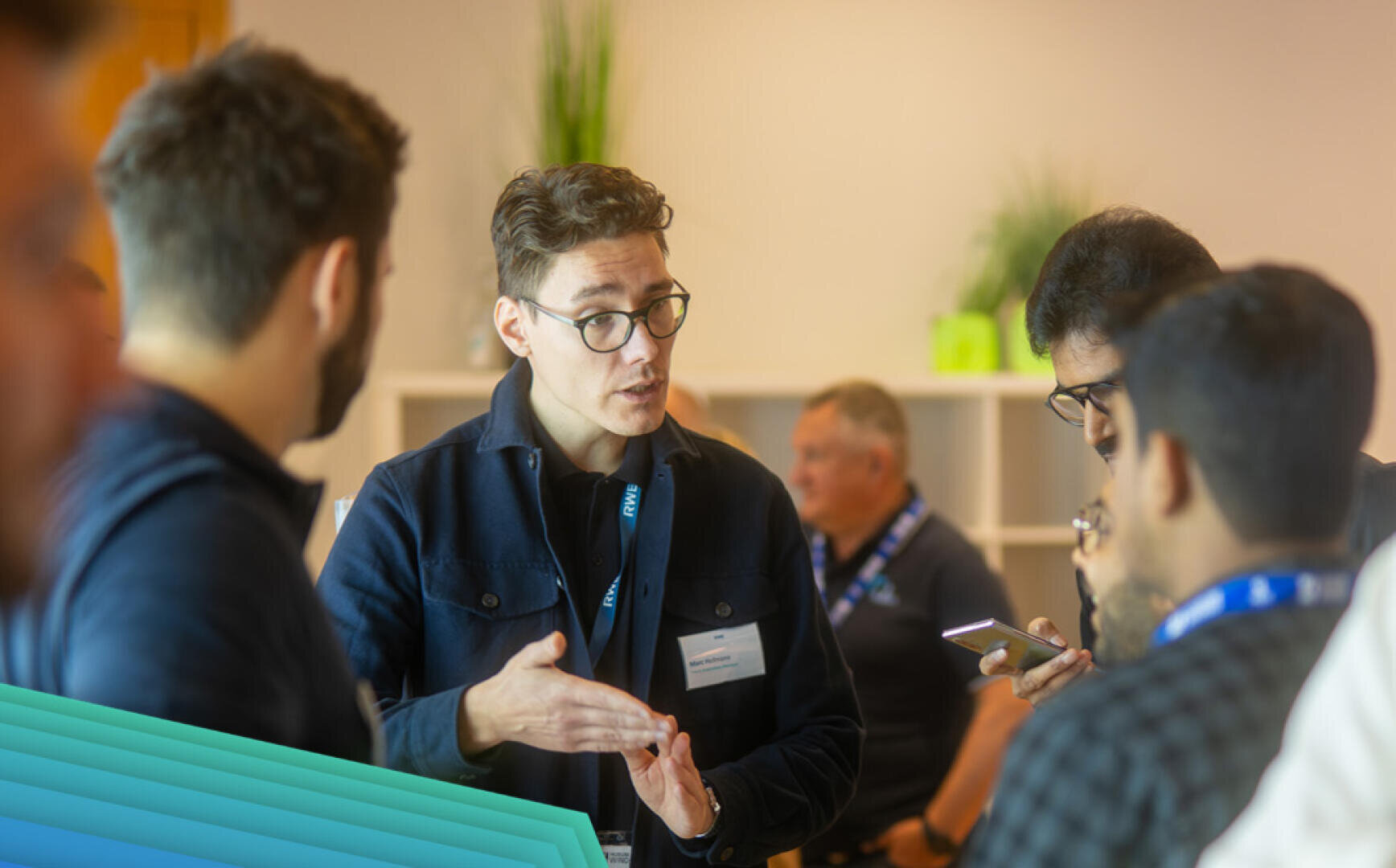 Eine Gruppe von Personen unterhält sich bei einer Veranstaltung in einem Gebäude. Eine Person, die eine Brille trägt, gestikuliert beim Sprechen, während die anderen zuhören und auf ihre Telefone schauen. Im Hintergrund sind Regale und Pflanzen zu sehen.