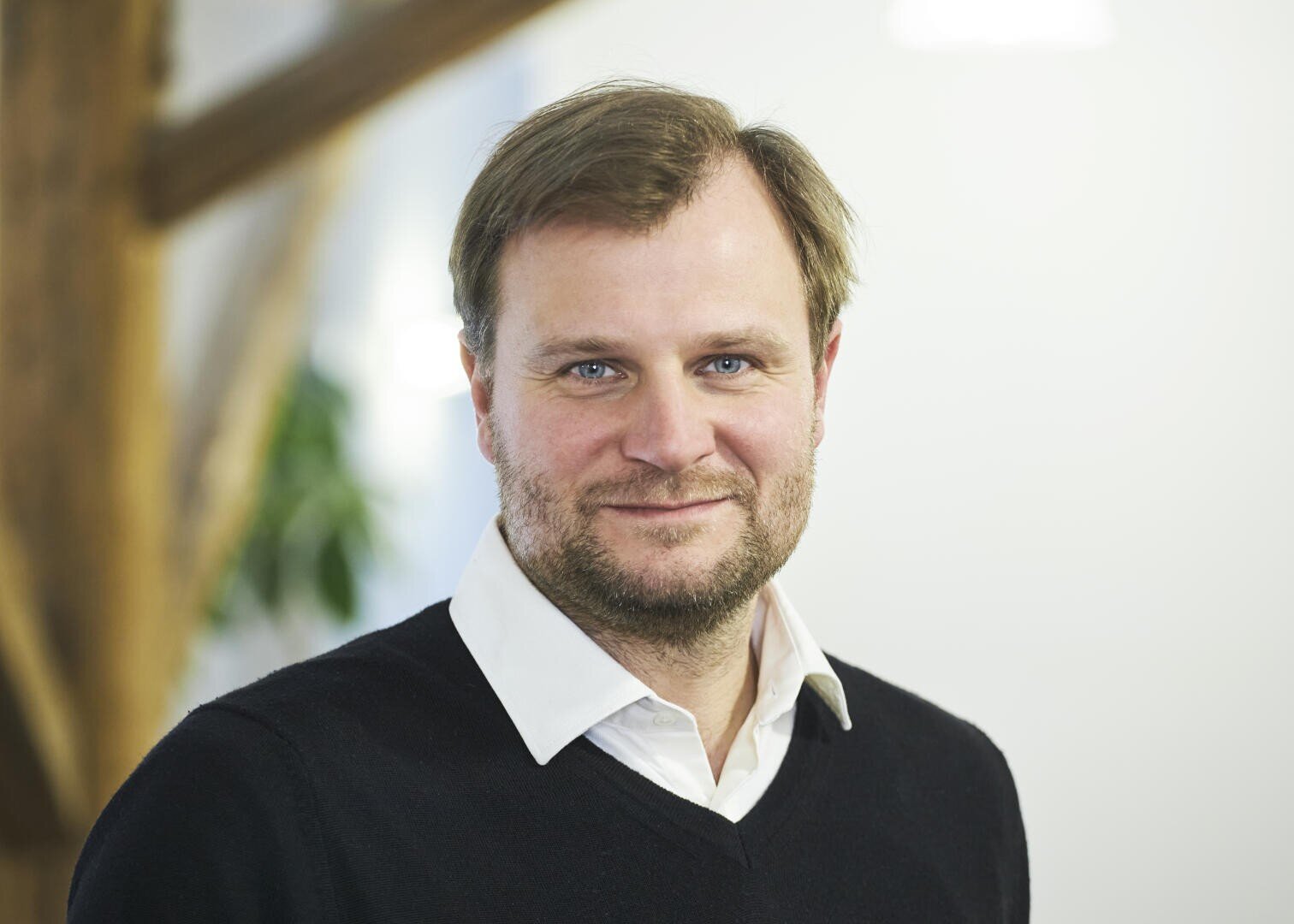 A man with light brown hair and a beard, wearing a white collared shirt and a black sweater, stands indoors with a soft smile. The background is blurred, showing some greenery and wooden beams.