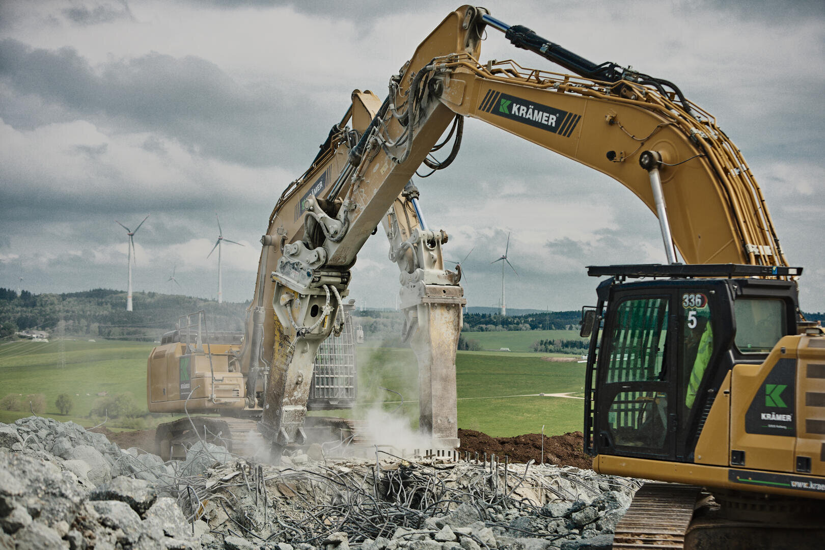 Two large yellow excavators remove concrete and rebar at a construction site. Dust swirls from the debris. In the background, wind turbines and green fields can be seen under a cloudy sky.