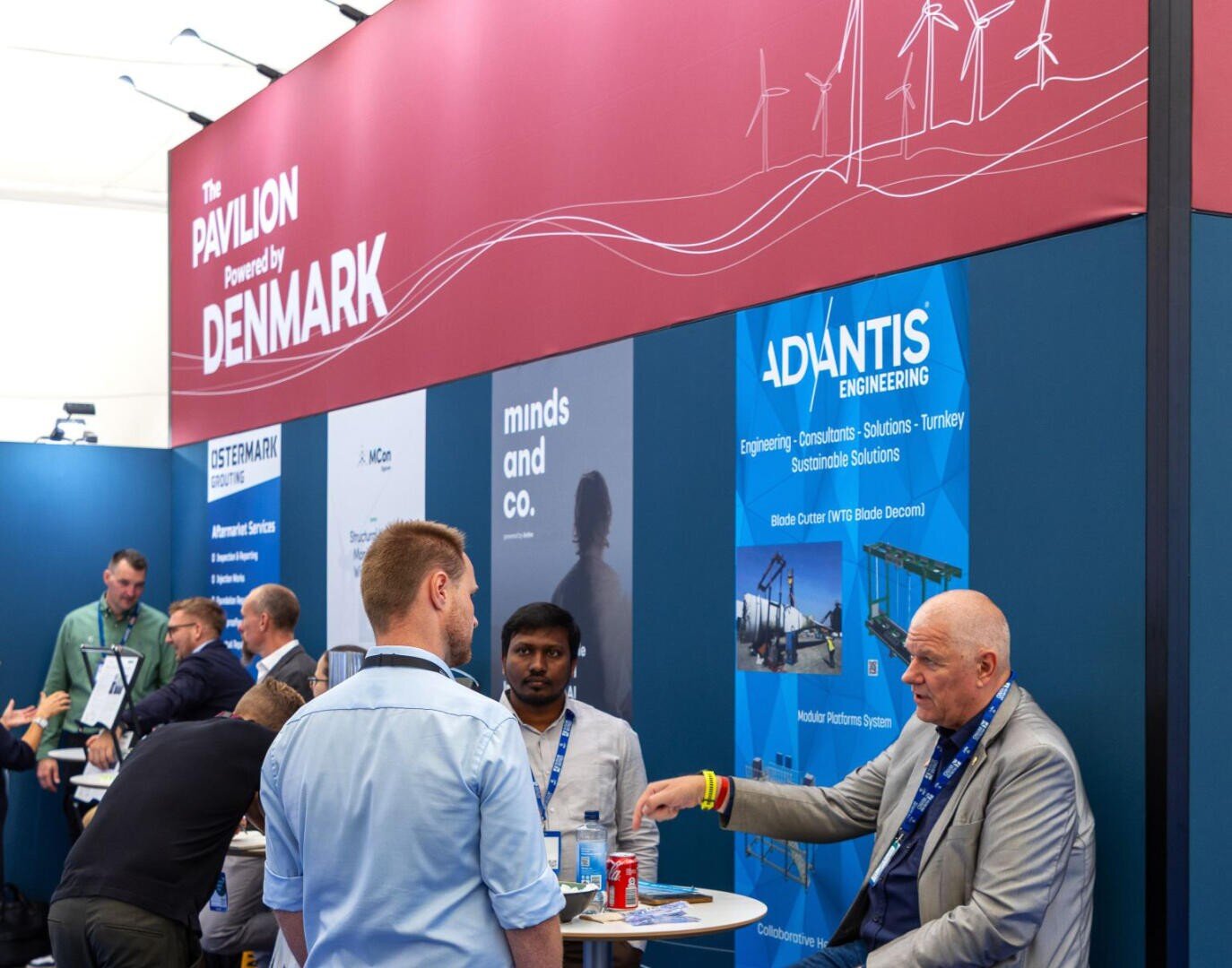 A group of people are talking at an exhibition stand under a red sign reading The Pavilion Powered by Denmark. Advertising banners and information material can be seen in the background.