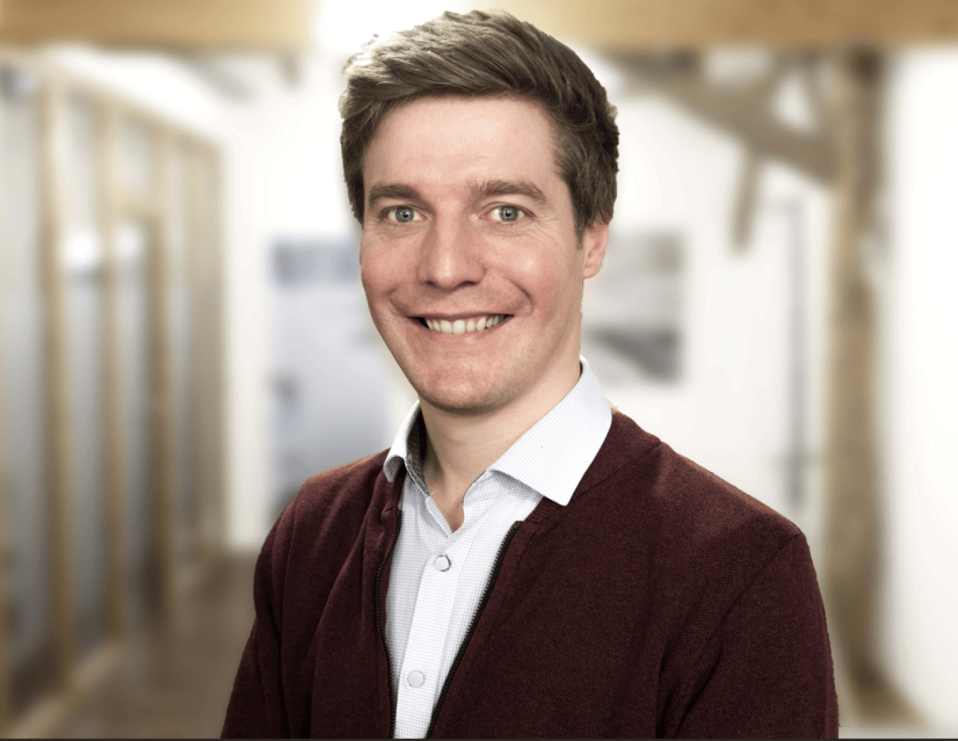 Ein lächelnder Mann mit kurzen braunen Haaren, der einen kastanienbraunen Pullover über einem weißen Hemd mit Kragen trägt, steht drinnen in einer unscharfen Büro- oder Flurumgebung.