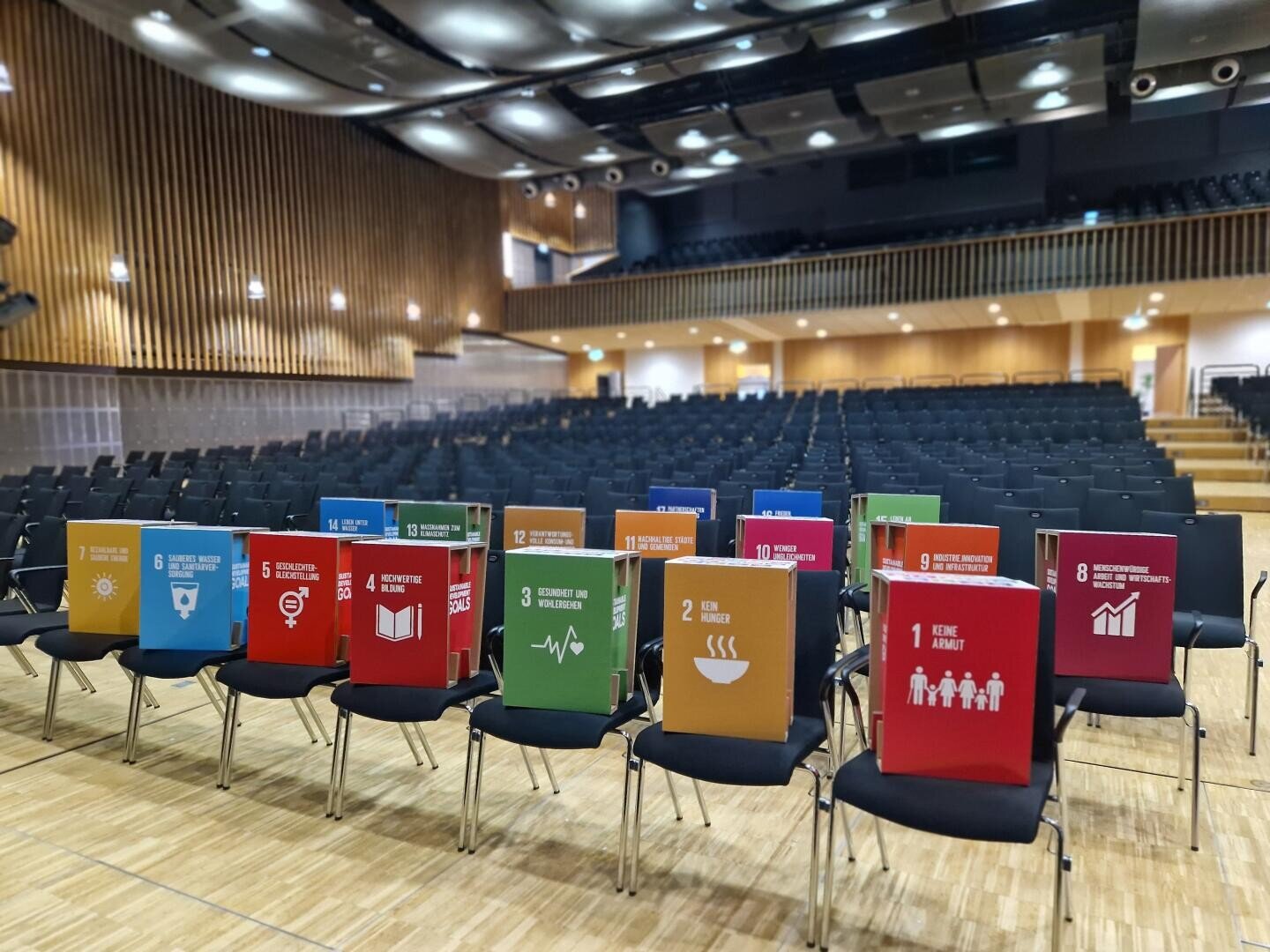 An empty auditorium with rows of chairs, some front seats marked with colorful posters displaying icons and numbers representing the United Nations Sustainable Development Goals.