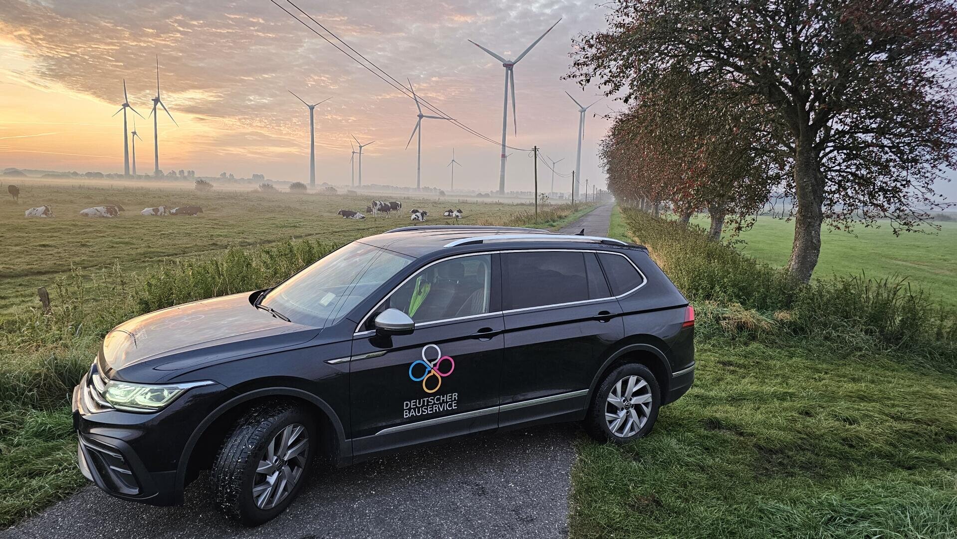 Ein schwarzer SUV mit dem Logo des Deutschen EnergieService parkt am grasbewachsenen Straßenrand in der Nähe einer Kuhweide. Im Hintergrund sind Windräder und Stromleitungen zu sehen, unter einem morgendlichen Himmel mit orangefarbenen Wolken.