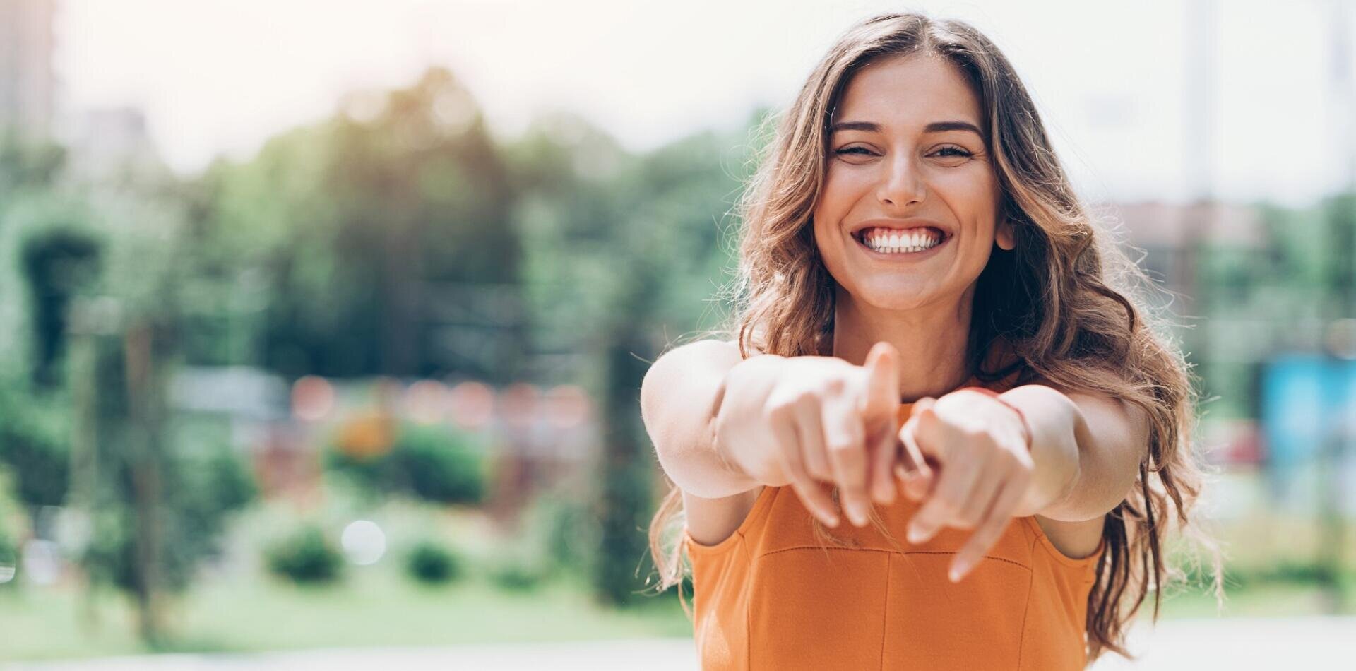 A smiling young woman with long wavy hair points toward the camera with both hands, standing outdoors on a sunny day with greenery in the background.