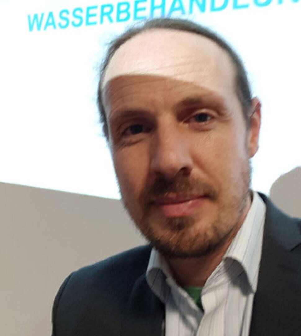 A man with light skin, a beard, and long hair tied back is wearing a collared shirt and suit jacket, standing indoors. The background shows part of a projected word in German: WASSERBEHANDLUNG.