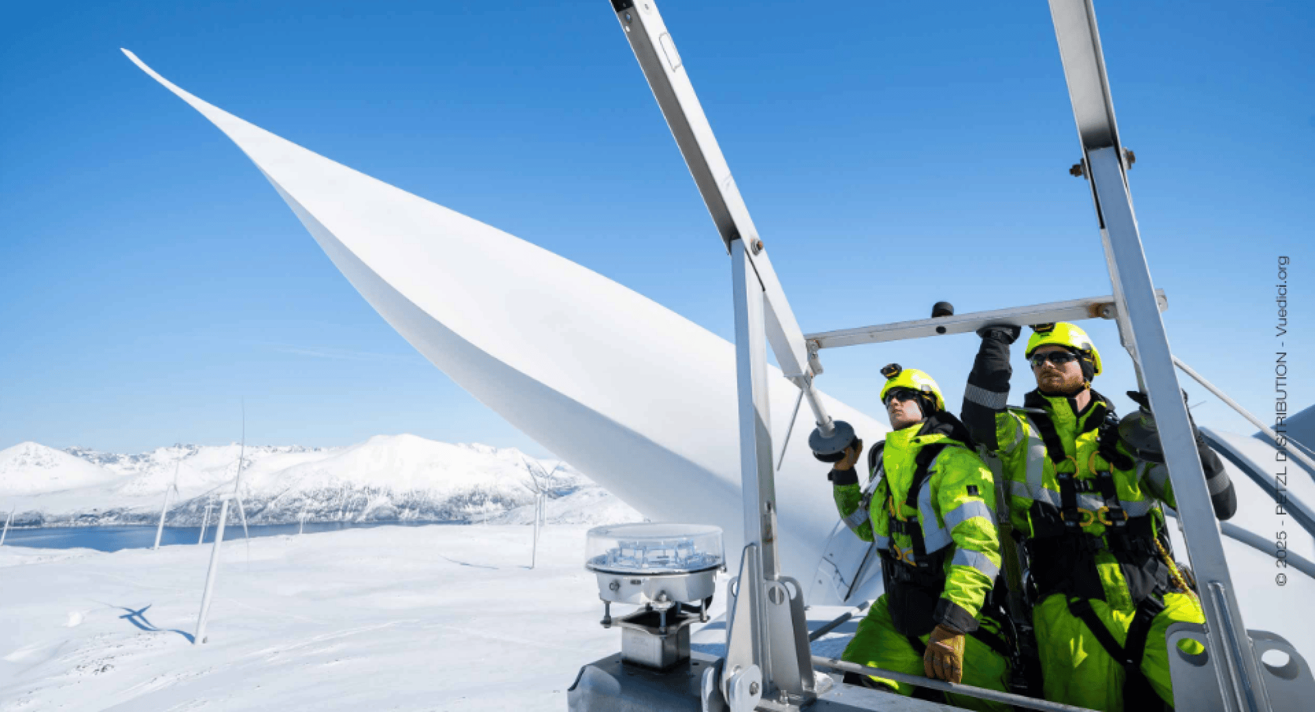 Zwei Arbeiter in heller Schutzkleidung inspizieren eine Windkraftanlage in einer verschneiten Landschaft, in der Berge und andere Windkraftanlagen unter einem klaren blauen Himmel zu sehen sind.
