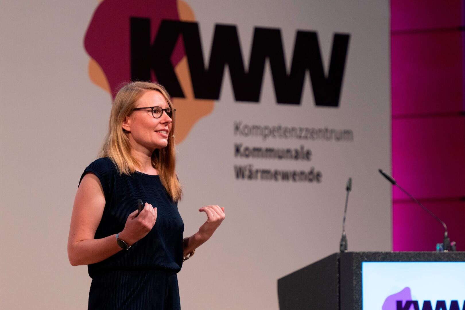 A woman with blonde hair and glasses speaks on stage in front of a podium with microphones. Behind her is a large sign with the inscription KWW Kompetenzzentrum Kommunale Wärmewende.