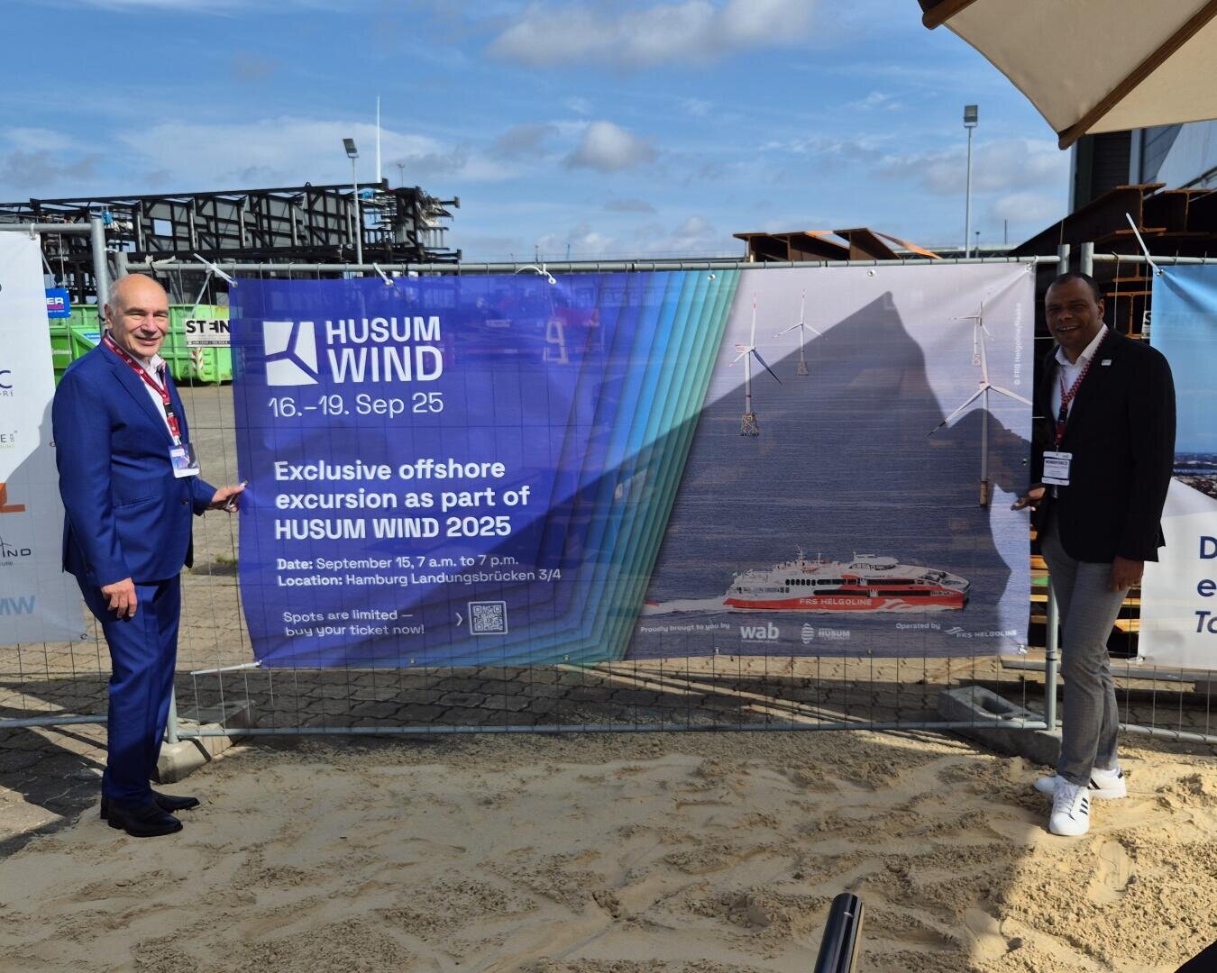 Zwei Männer in Geschäftskleidung stehen im Sand an einem Zaun und zeigen auf ein großes Banner, das für die Offshore-Exkursion Husum Wind 2025 wirbt. Auf dem Banner sind Windturbinen, ein Schiff und Veranstaltungshinweise zu sehen.