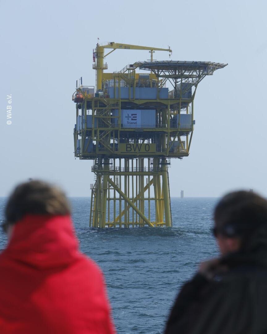 Zwei Personen in Jacken betrachten eine große gelbe Offshore-Plattform, die unter einem dunstigen Himmel im Meer steht. Auf der Plattform befinden sich Maschinen und ein kleines Gebäude. Das Wasser ist ruhig.