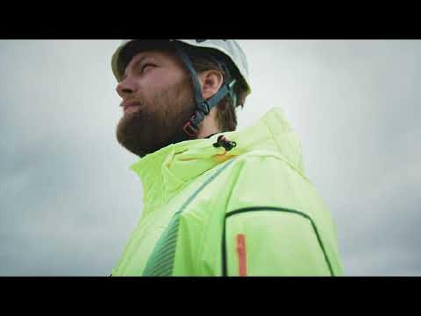 Eine bärtige Person mit weißem Schutzhelm und leuchtend gelber Warnweste blickt vor einem bewölkten Himmel nachdenklich in die Ferne.