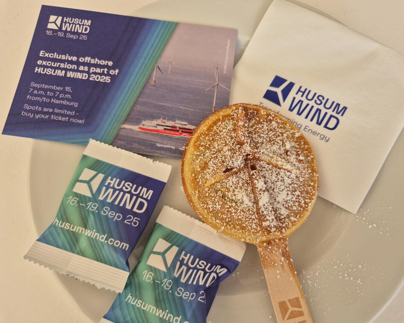 Ein Teller mit einer Puderzuckertorte am Holzstab, zwei in HUSUM WIND verpackte Bonbons, eine gebrandete Serviette und ein Flyer für eine Offshore-Exkursion im Rahmen von HUSUM WIND 2025 mit Windrädern und einem Schiff.