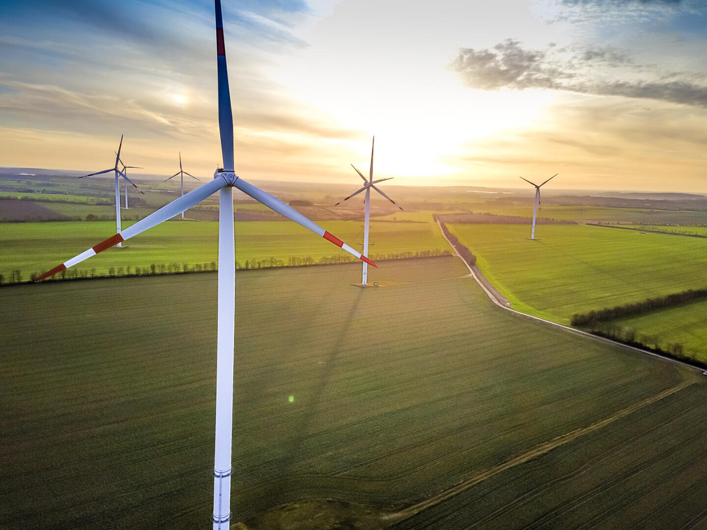 Aerial view of wind turbines on vast green fields at sunset, when the sky is partly cloudy and soft sunlight illuminates the landscape.