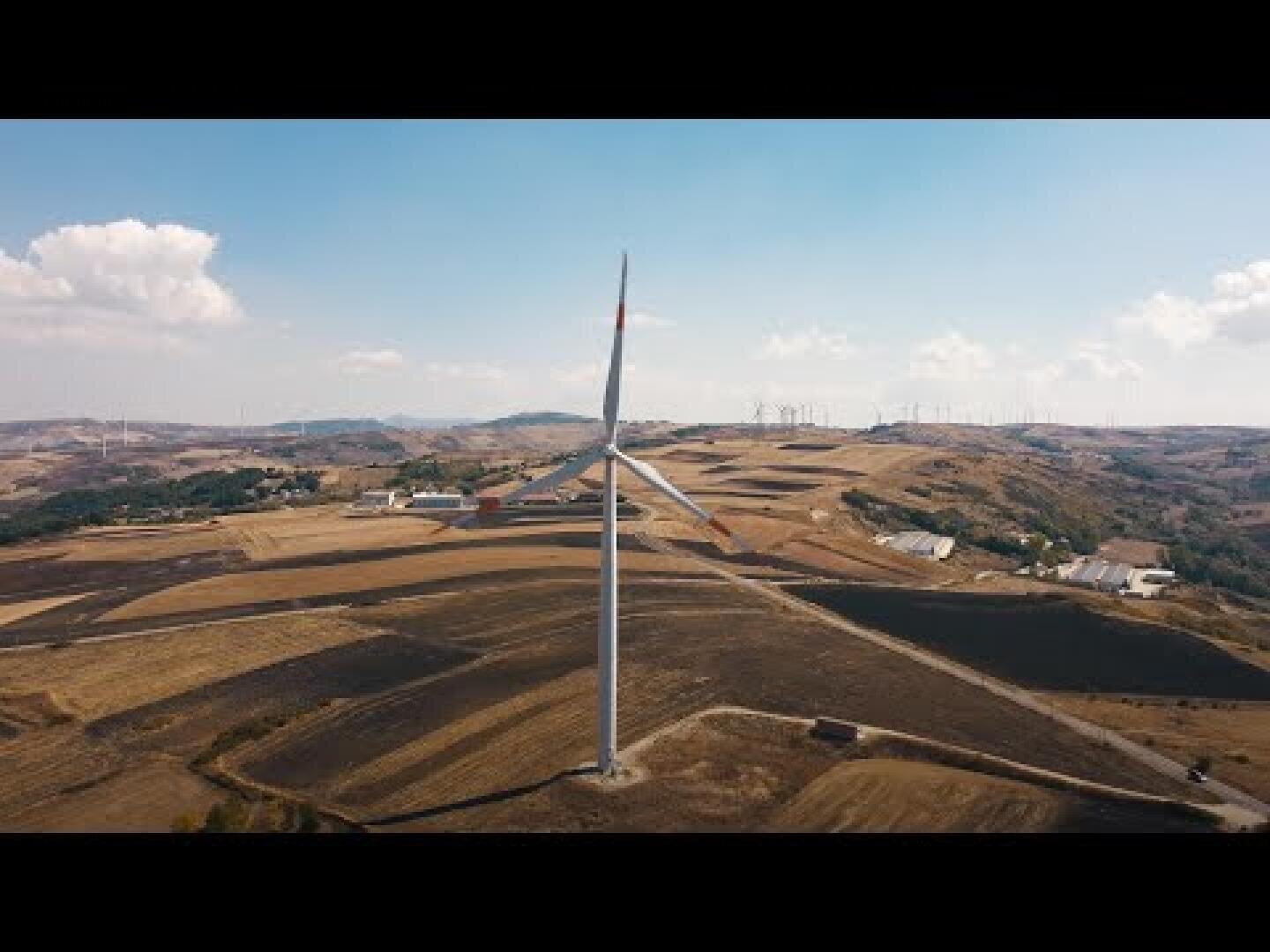 Eine Windkraftanlage steht inmitten einer trockenen, hügeligen Landschaft mit verstreuten Gebäuden und Feldern unter einem teilweise bewölkten Himmel. In der Ferne sind weitere Windkraftanlagen zu sehen.