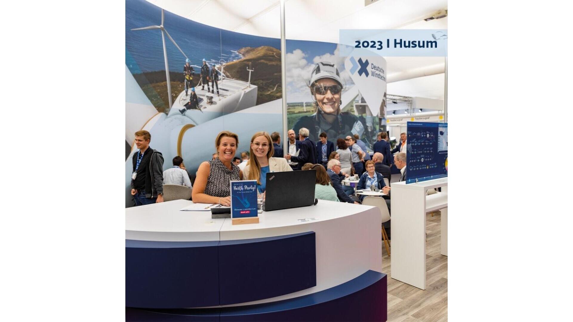 Three women smile at a reception desk with a laptop at an indoor expo. Behind them, people mingle near large wind turbine images and a 2023 | Husum sign. The setting is bright and busy.