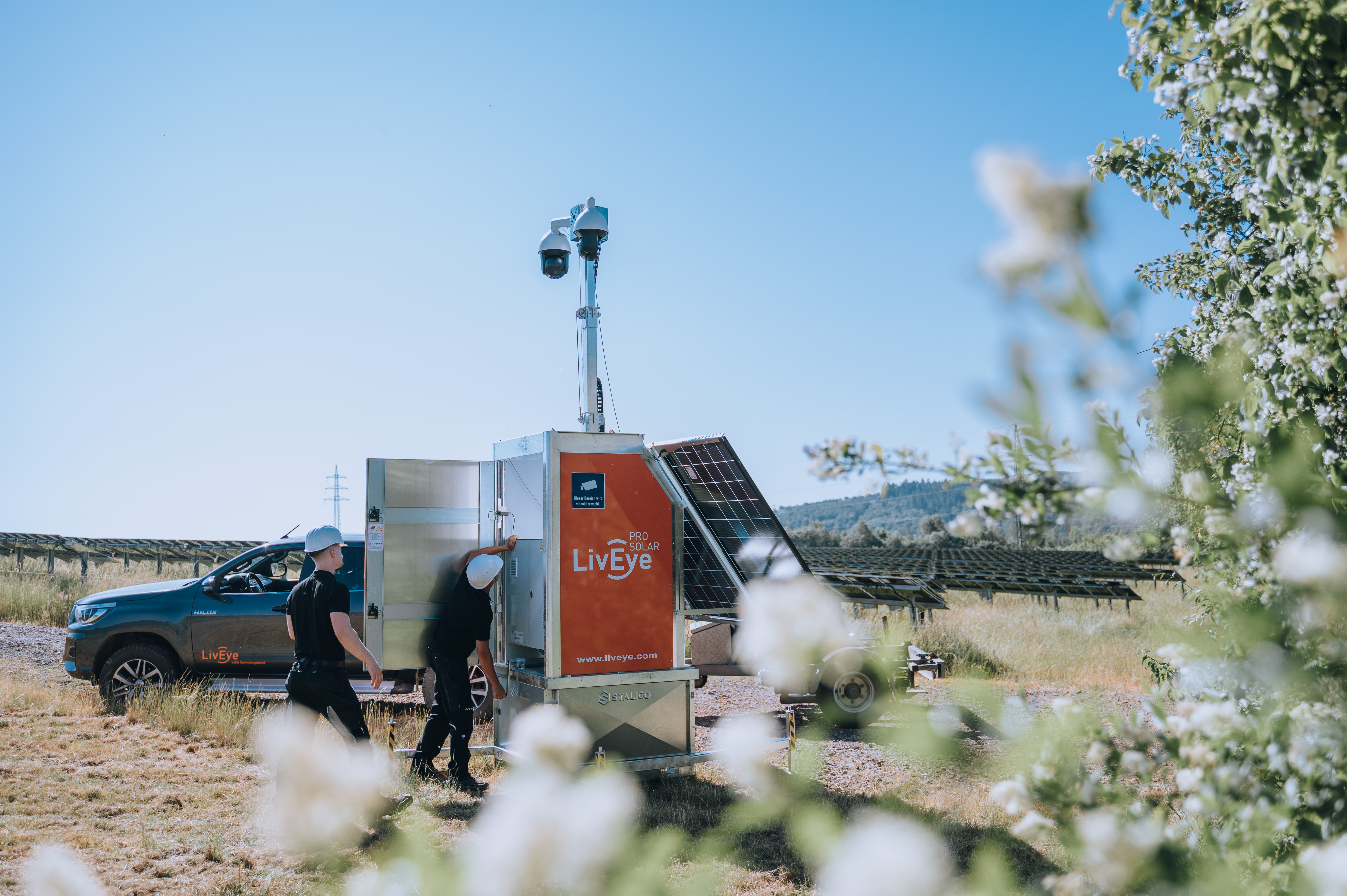 Two people in hard hats inspect a mobile surveillance unit with solar panels labeled Live Eye in a grassy field, near a black pickup truck, with power lines and solar panels in the background.