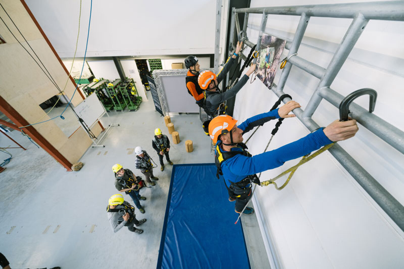 Mehrere Personen mit Helmen und Schutzausrüstung klettern in einem Innenraum auf eine Metallkonstruktion, während andere von unten zusehen. Zu Sicherheitszwecken ist darunter eine blaue Matte ausgelegt. Der Ort scheint eine Trainingsanlage zu sein.